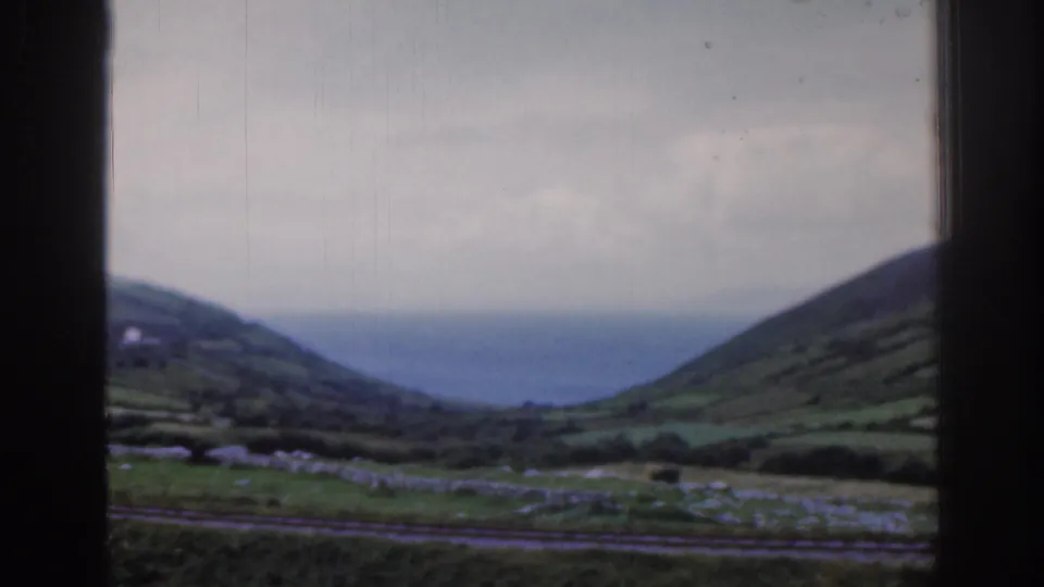 1961 Ireland 8mm frame showing rolling green hills, stone walls, rail tracks, and distant water seen through a dark train window.