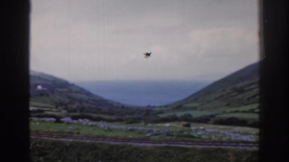 1961 Ireland train-window frame with green hills, stone-walled fields, small buildings, and distant coastal water under overcast light.