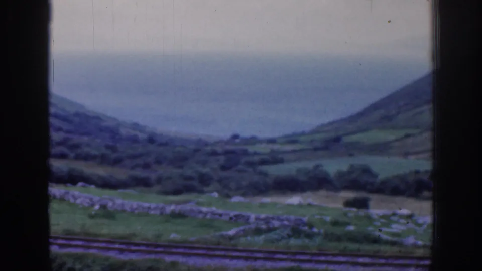 Soft 8mm frame from a moving train in Ireland, showing tracks, stone walls, fields, hills, and water softened by motion blur.