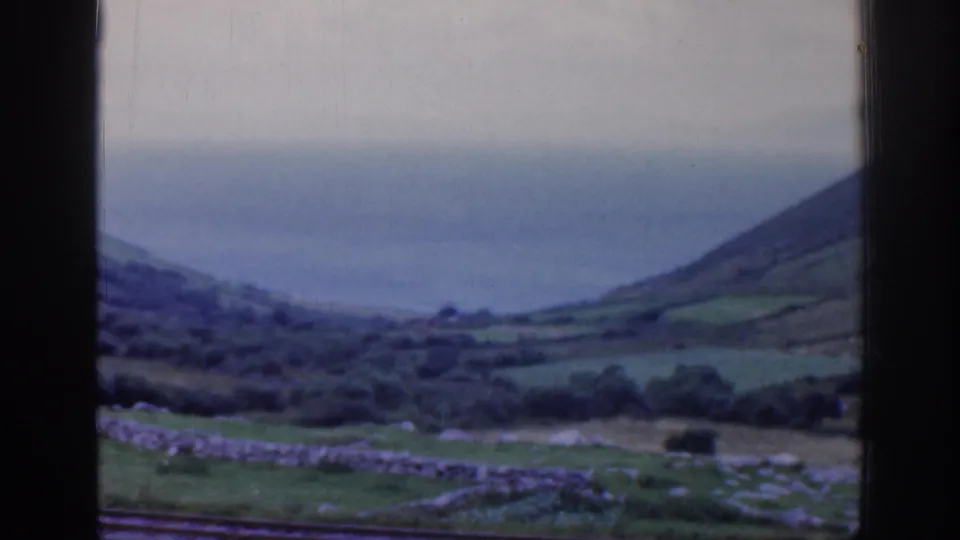 1961 Ireland 8mm train-window view of a hillside above stone-walled fields and tracks, with distant water under a cloudy sky.
