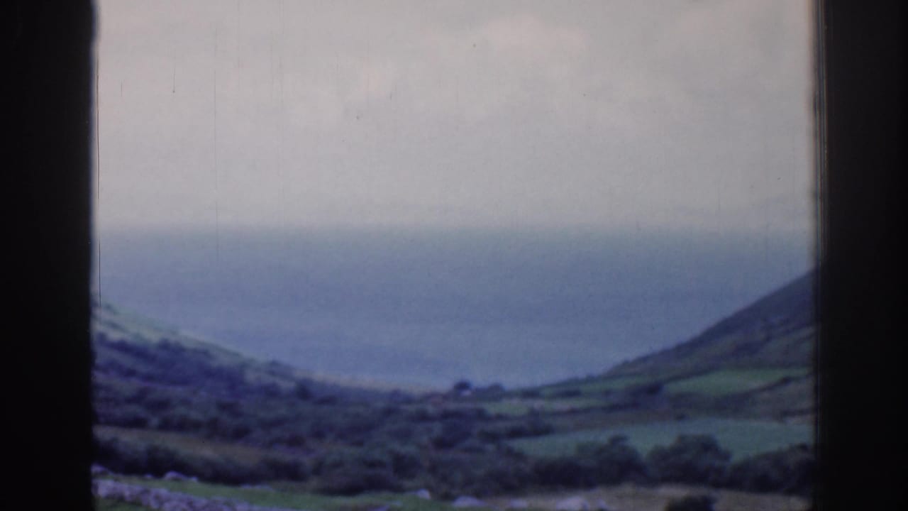 Rolling coastal hillside, stone walls, and sea behind a window frame.