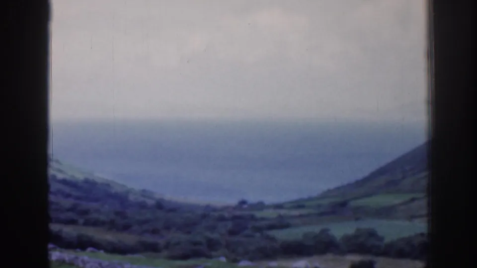 Soft 8mm frame of Irish coastal hills, stone-walled fields, and distant sea seen through the window opening.