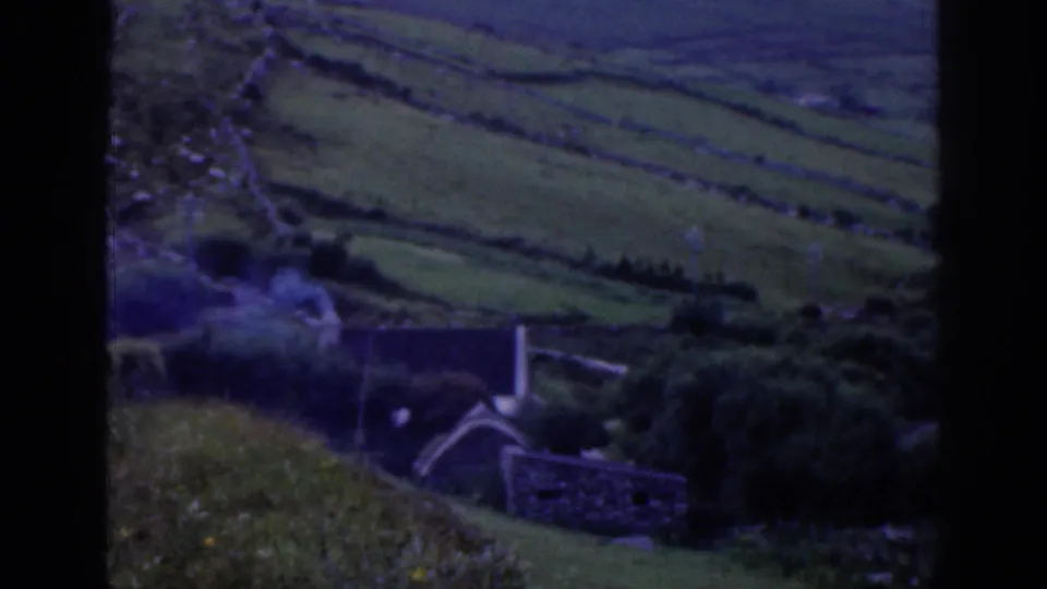 1961 Ireland home-movie frame showing stone-walled green fields and a small rural cottage in the valley.