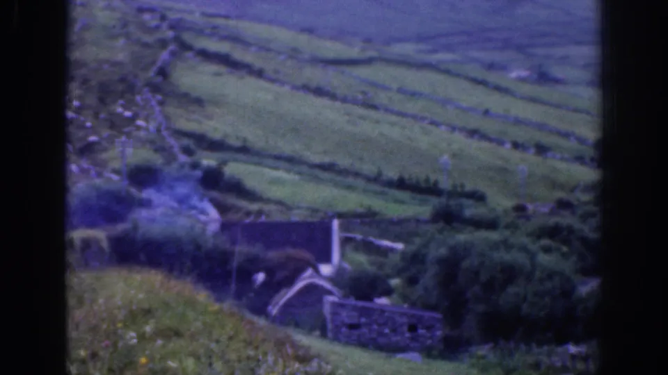 Vintage 8mm view of rural Ireland with a small cottage, trees, stone walls, and sloping green fields.