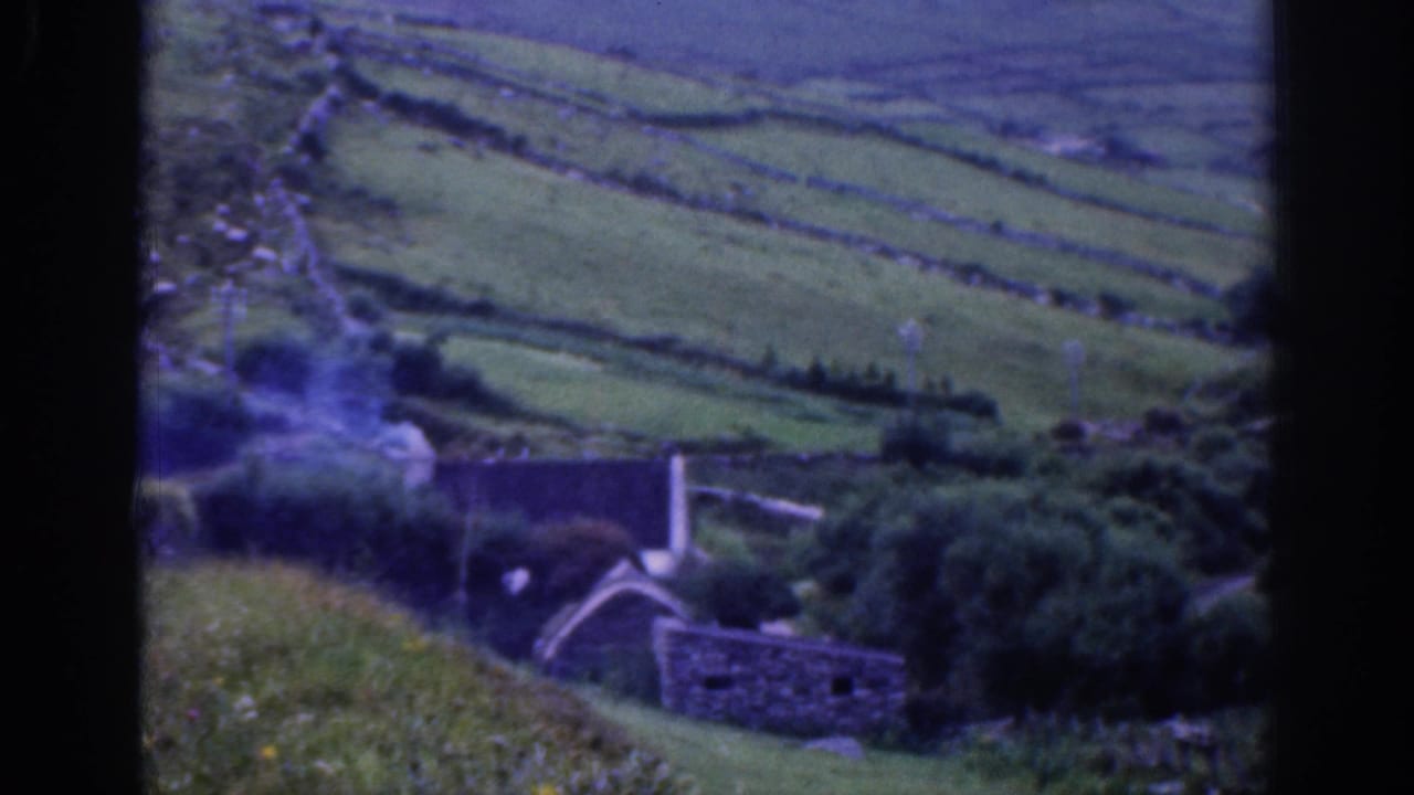 1961 Ireland home-movie frame showing terraced green hills, dry-stone walls, and a small stone structure in soft 8mm color.