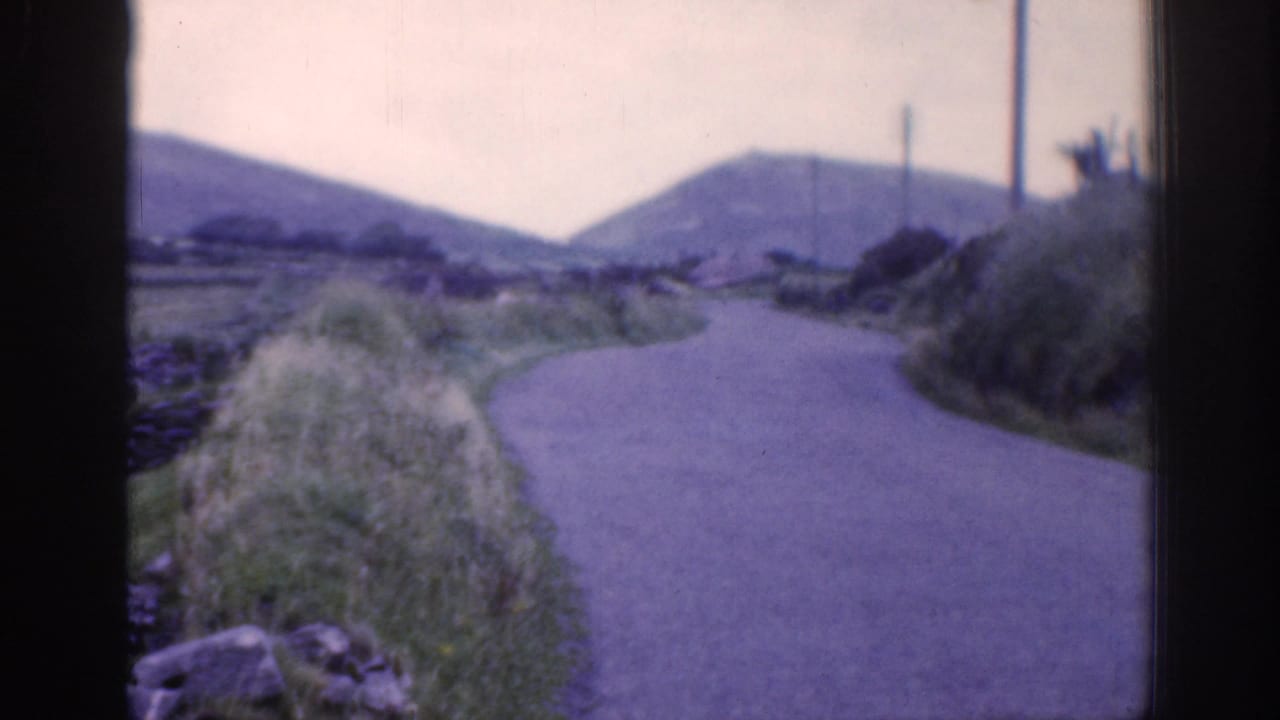 1961 Ireland countryside road curves past grassy banks and stone walls toward low hills under a pale sky.