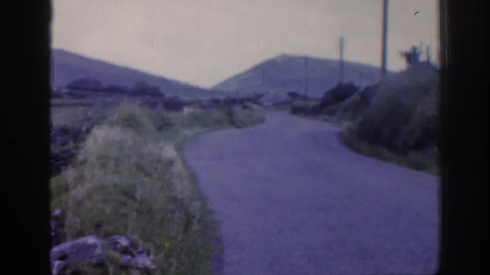 Soft 8mm view of a rural Irish road bending between grass, stone walls, utility poles, and distant hills in 1961.
