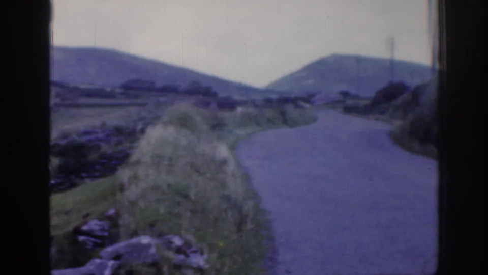 1961 Ireland roadscape with a curving paved lane, stone walls, grassy verge, and low hills seen through aged 8mm film.
