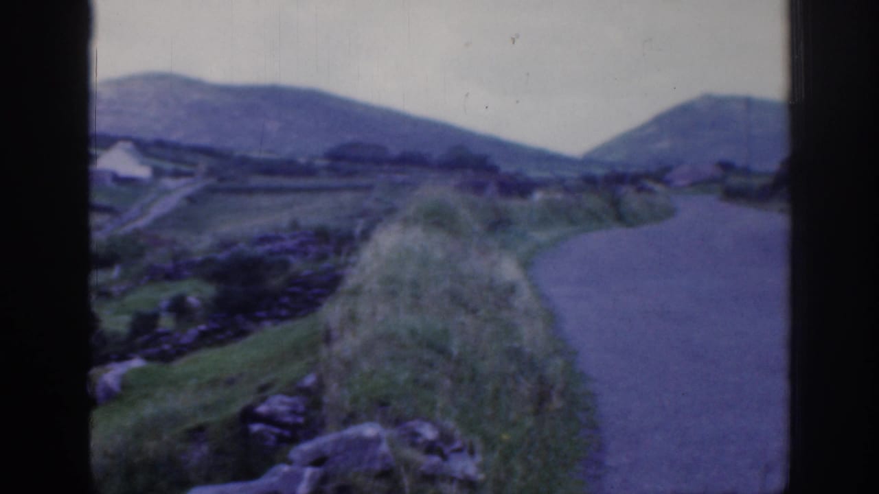 1961 Ireland home-movie frame showing a narrow rural road, rolling green hills, stone walls, and foreground rocks.
