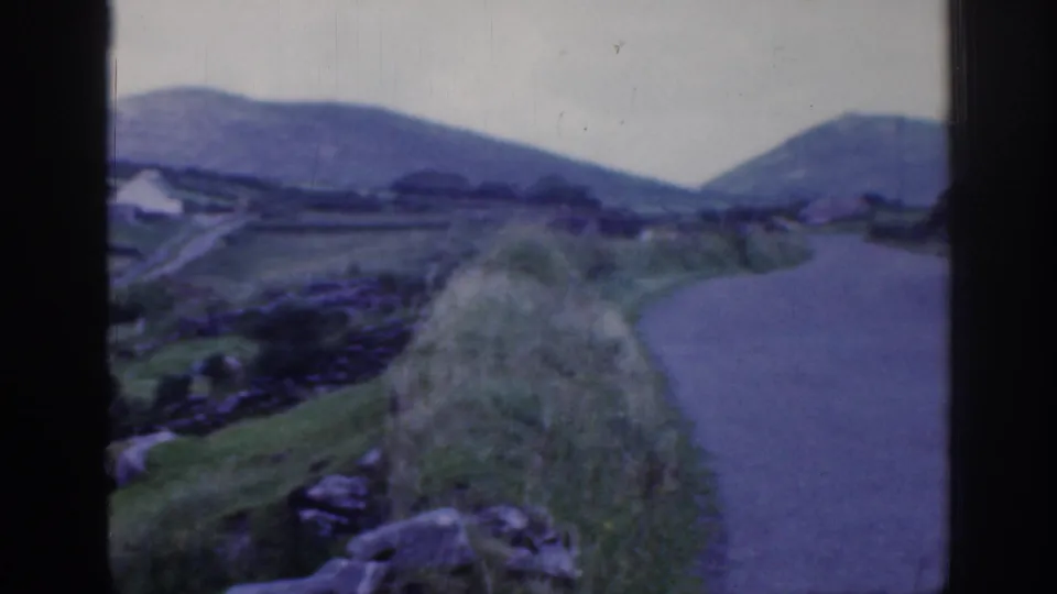 1961 Ireland home-movie frame showing a narrow rural road, rolling green hills, stone walls, and foreground rocks.