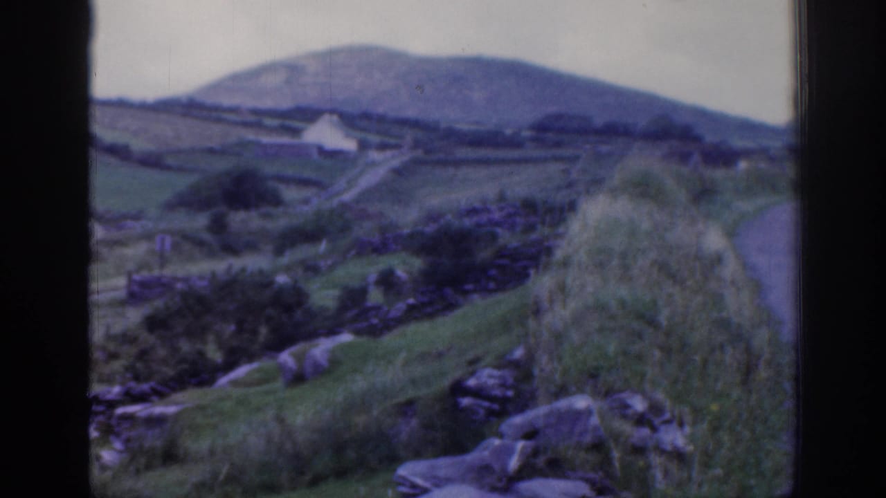 Green hillside landscape with stone walls, rocks, a small white building, and distant hills.