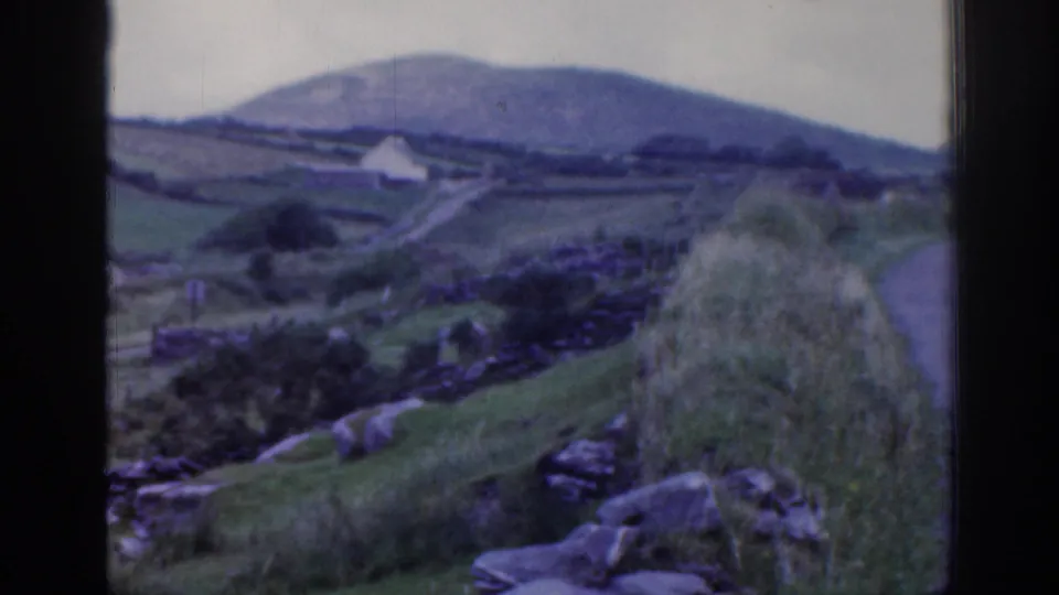 1961 Ireland countryside view with stone-walled fields, rocks, grassy slopes, a small white building, and distant hills.