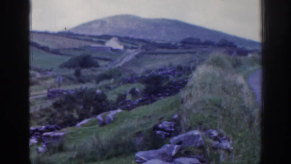 1961 Ireland home-movie frame of a grassy hillside, stone walls, rocks, a narrow road, and a small distant building.