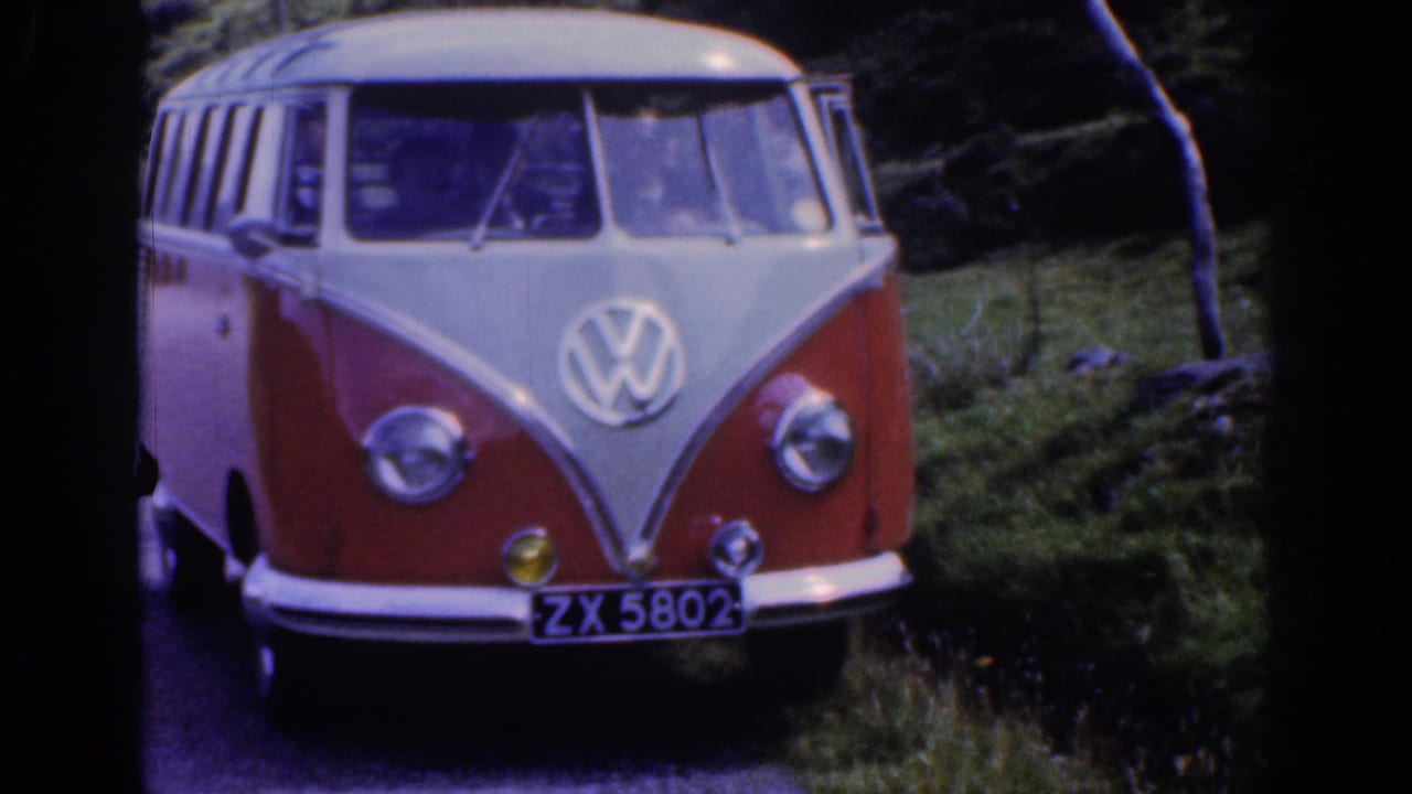 1961 Ireland home-movie frame showing a red and white Volkswagen camper close to camera on a green rural roadside.