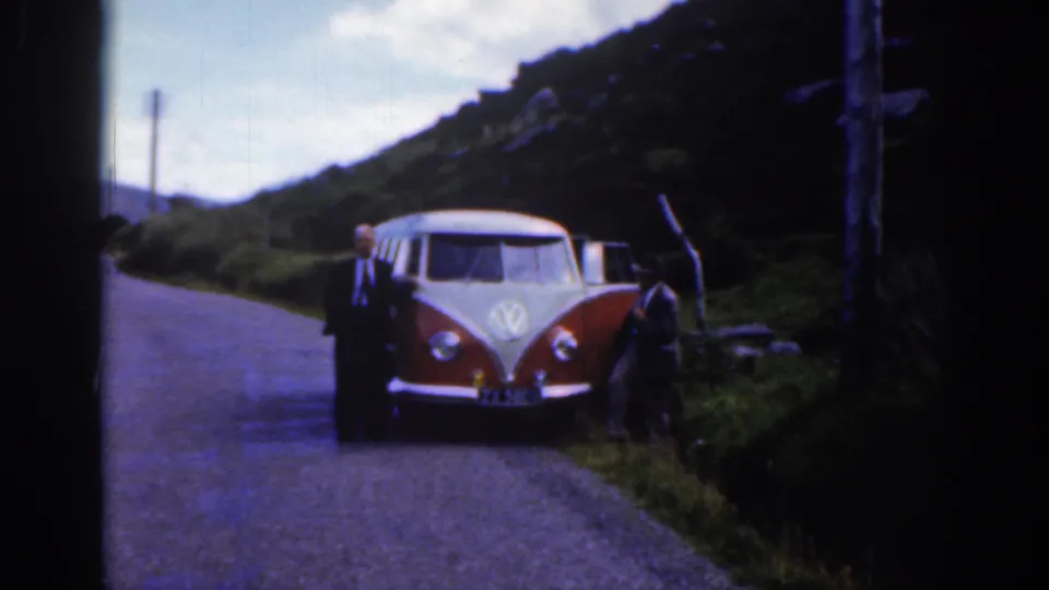 Wider 1961 Ireland roadside view with two suited men near the Volkswagen camper, hills, grass, and cloudy sky behind them.
