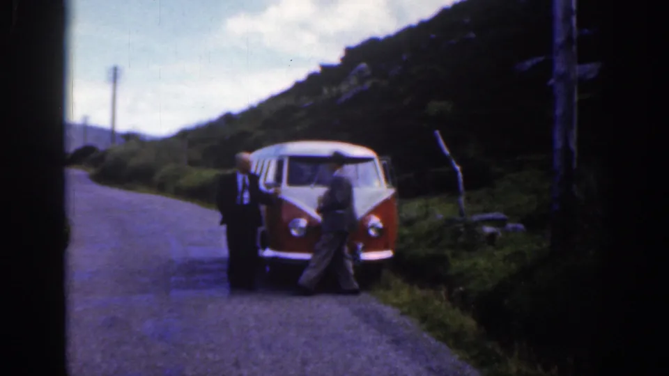 Wider home-movie frame of men beside the red and white Volkswagen camper on a rural Irish road with hills and grass visible.