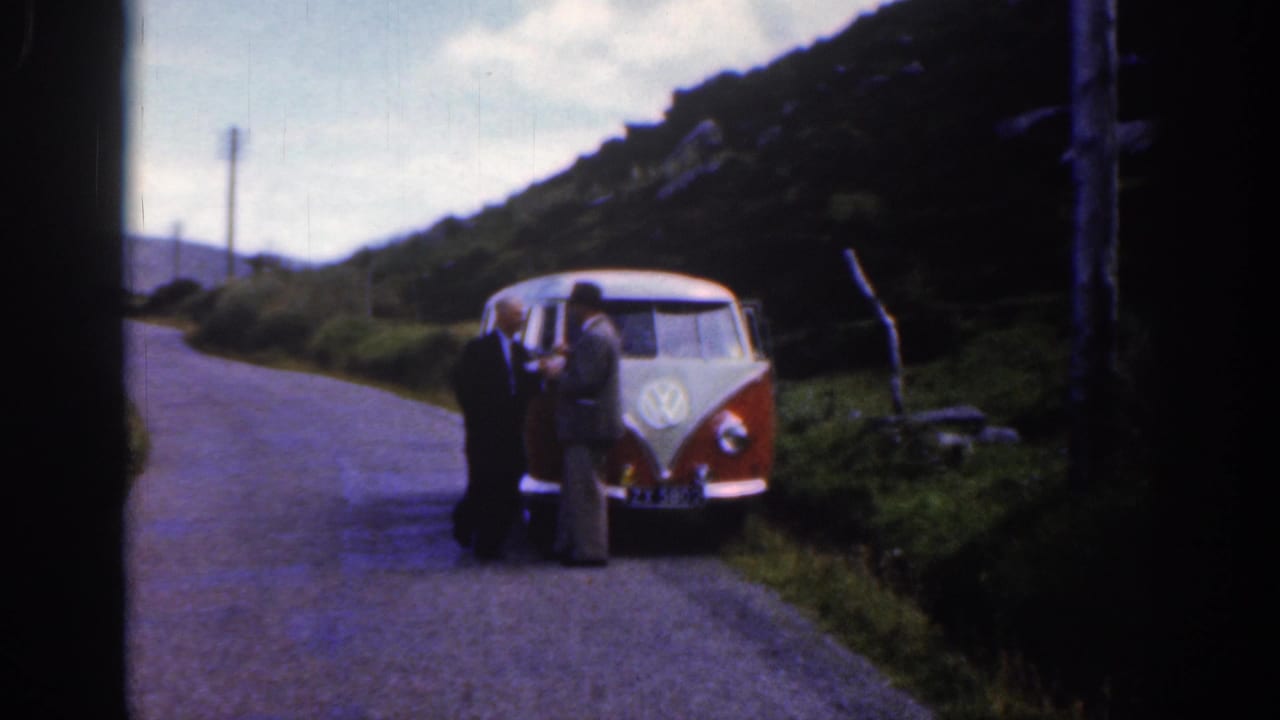 Two adult men stand at the front of a red and white Volkswagen bus on a narrow rural road in Ireland, 1961.