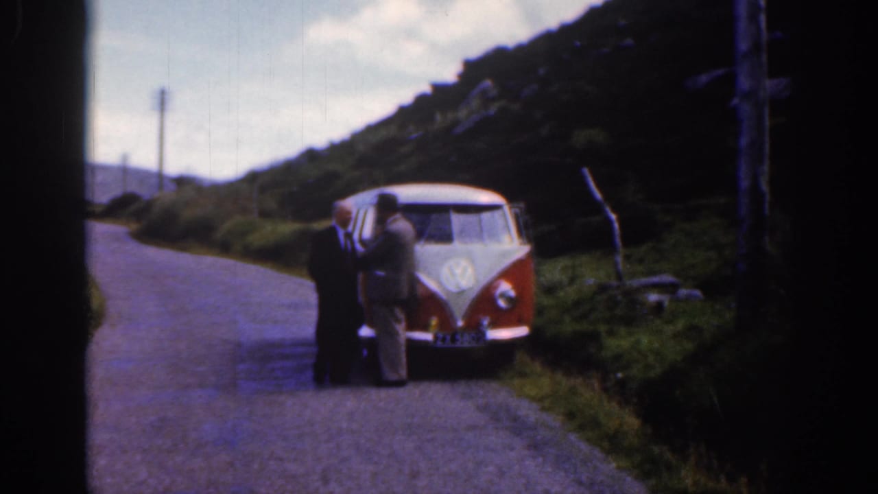 Two men in suits stand beside a red and white Volkswagen bus on an Irish rural road, with hills and roadside grass visible.