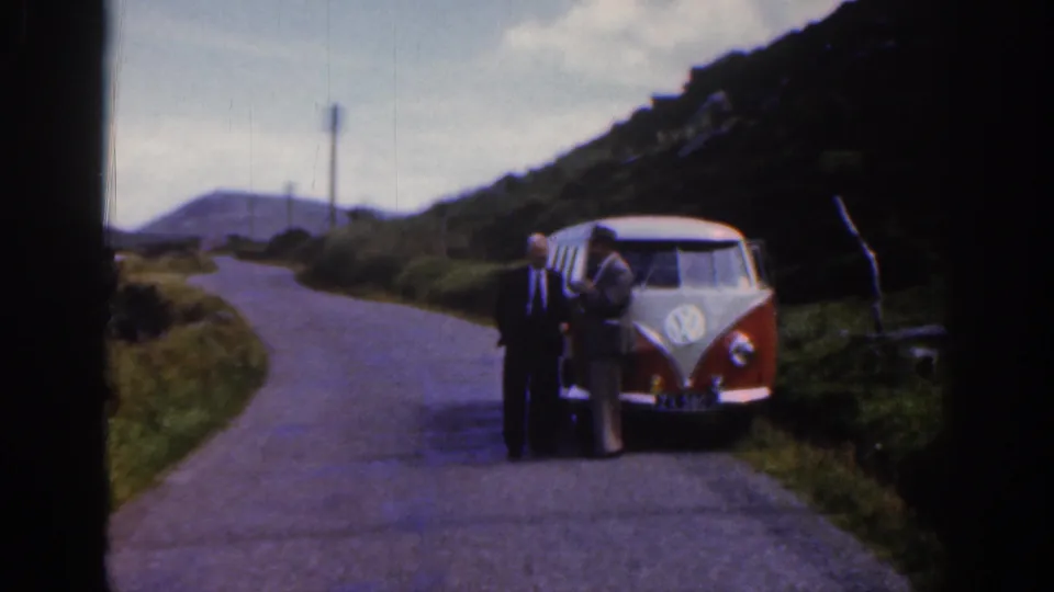 Two adult men stand by a parked Volkswagen bus on a curving rural road in Ireland, with hills and roadside vegetation nearby.