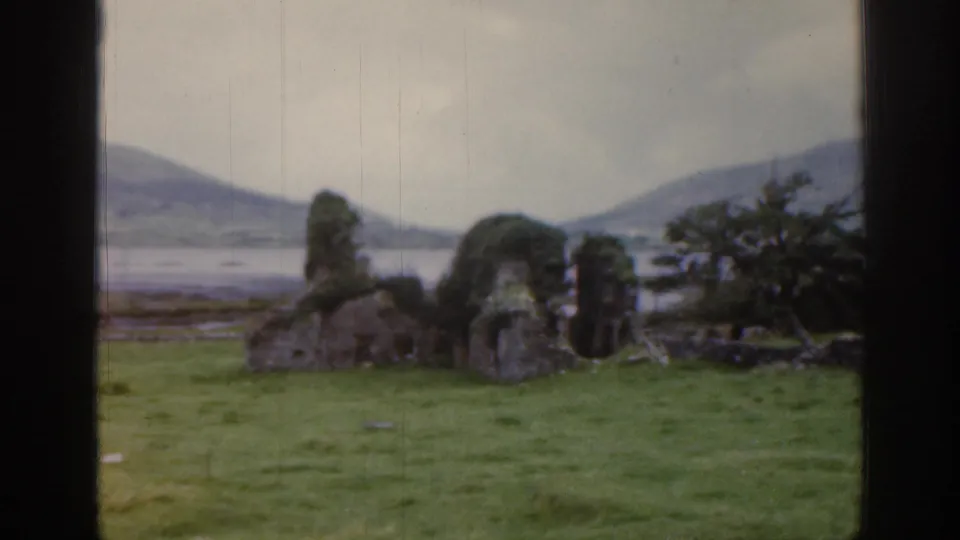 Mossy stone ruins sit in a green field near a lake or inlet, with low Irish hills in the background.