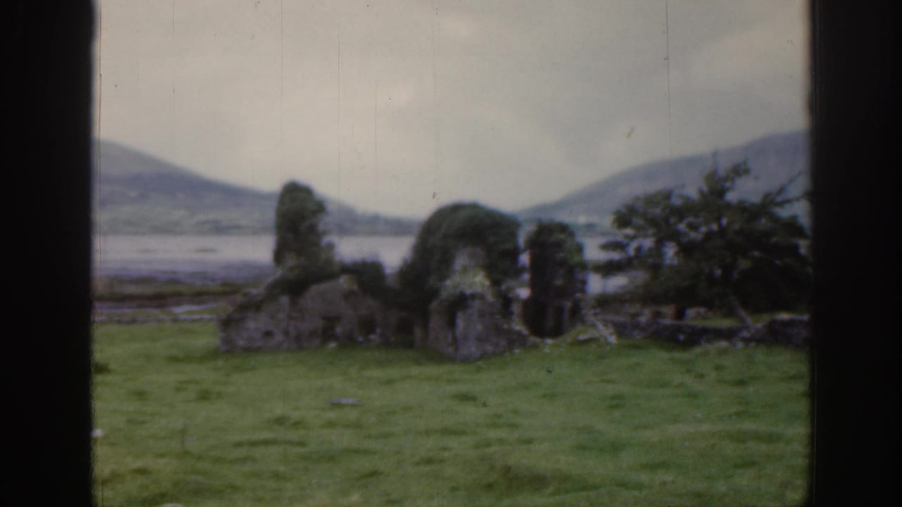1961 Ireland 8mm frame showing moss-covered stone ruins in a grassy field, with distant water, hills, and dark film borders.