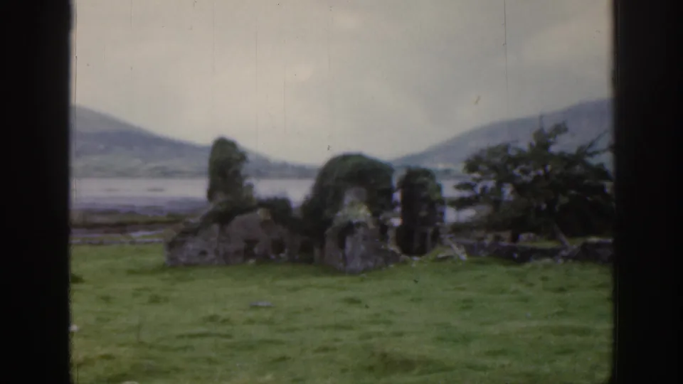 1961 Ireland 8mm frame showing moss-covered stone ruins in a grassy field, with distant water, hills, and dark film borders.