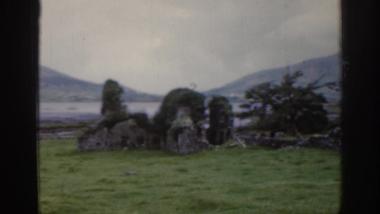 Stone ruins and shrubs in a green field with hills and water in the background.