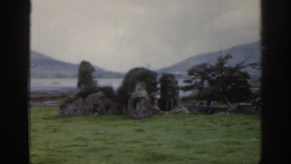 1961 Ireland home-movie frame with mossy stone remains, low walling, shrubs, distant water, and hills under an overcast sky.