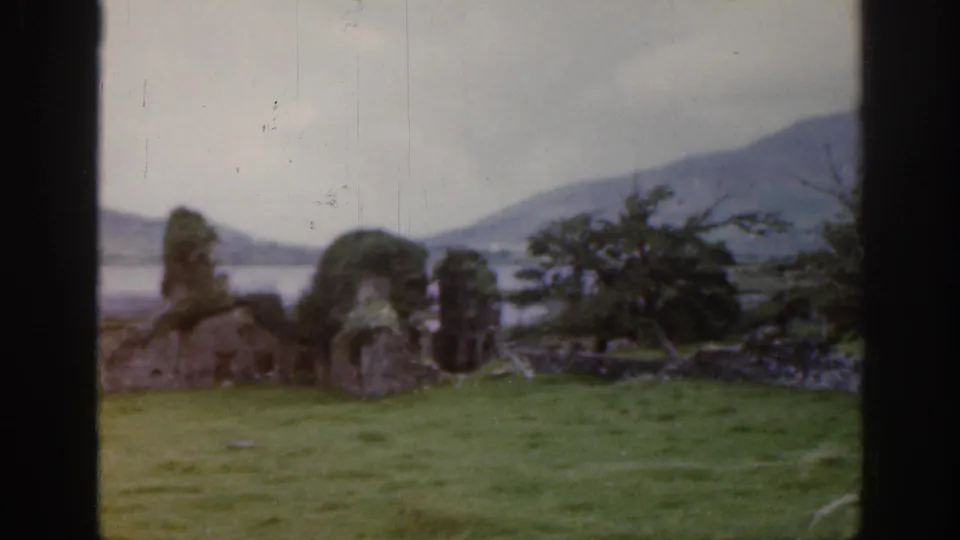 Soft 8mm frame from Ireland in 1961 showing moss-covered ruins, open grass, distant water, hills, and visible film-frame edges.