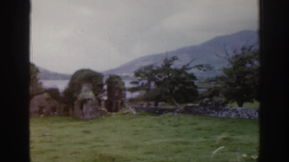 1961 Ireland 8mm frame of weathered stone ruins and a low wall in lush grass, backed by water, hills, and cloudy sky.