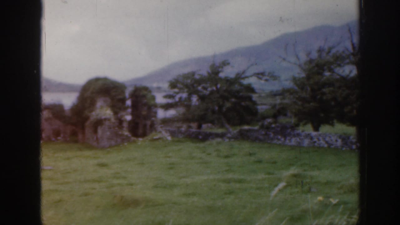 1961 Ireland home-movie frame of green countryside with trees, a stone wall, distant water, and hazy hills.
