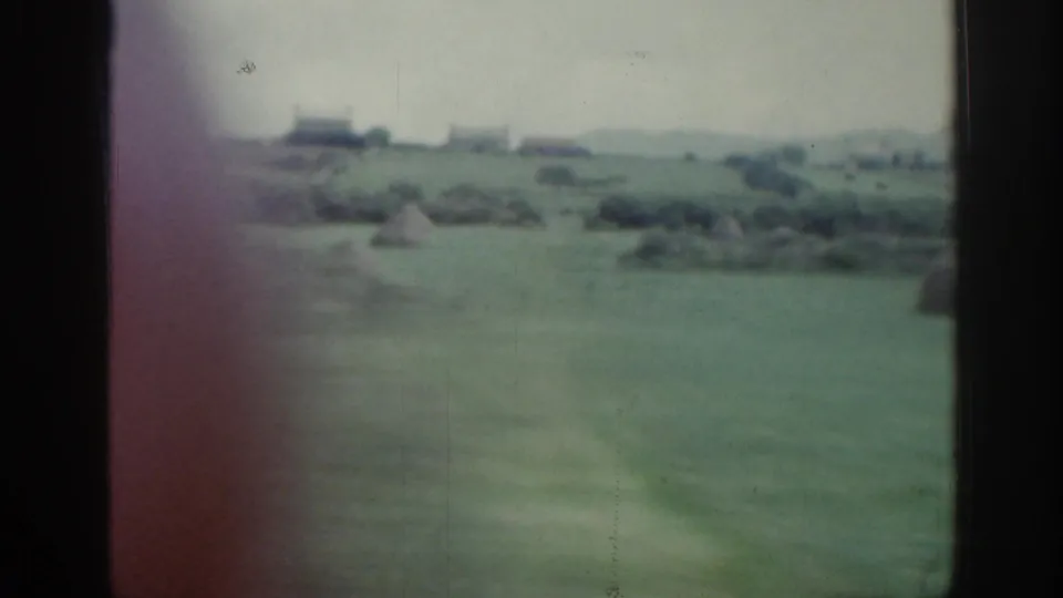 1961 Ireland countryside frame showing an open green field, haystacks, and distant low buildings under a pale sky.