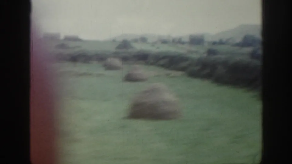 Soft 8mm frame of Irish countryside with haystacks in a green field, hedges, distant buildings, and low hills.
