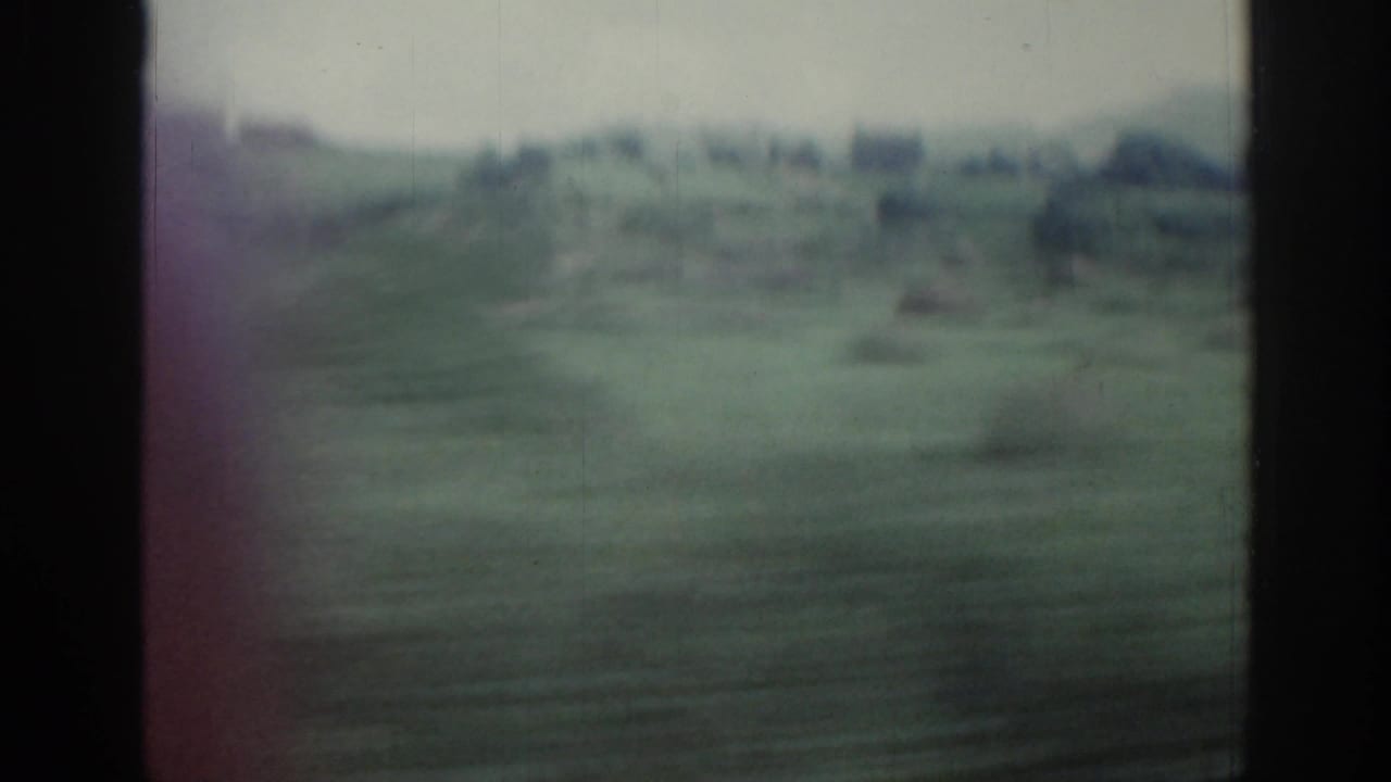1961 Ireland 8mm frame showing blurred fields and distant low buildings through dark train-window bars.