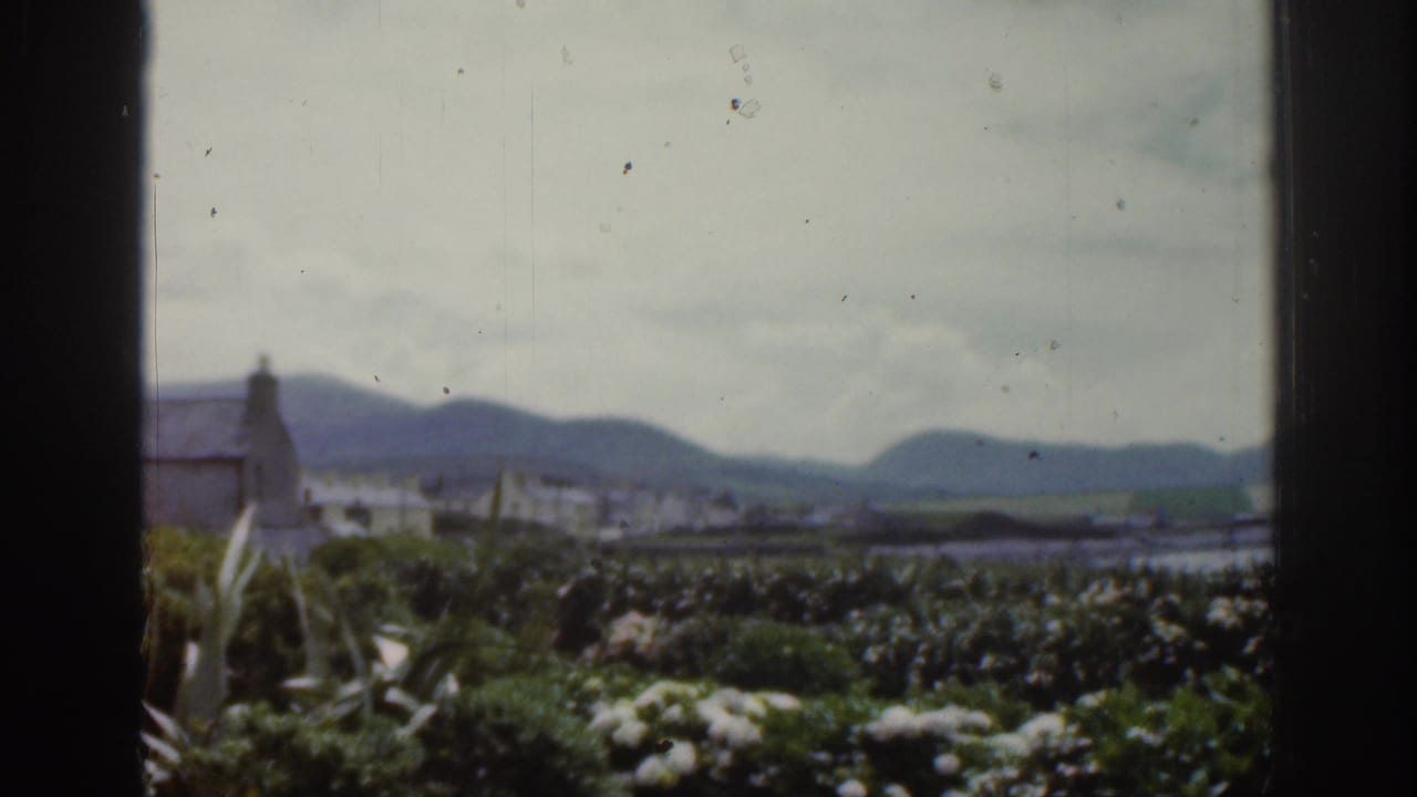 1961 Ireland home-movie view through a window toward a coastal town, green foreground, water, and low distant mountains.