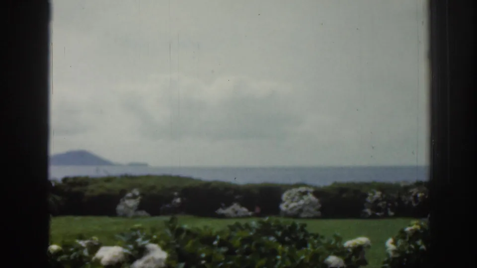 1961 Ireland coastal view through a window, looking past lawn and white shrubs toward open water and low mountains.