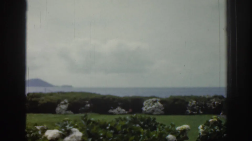 1961 Ireland 8mm frame showing a green garden and white flowering shrubs through window frames, with water and distant hills beyond.