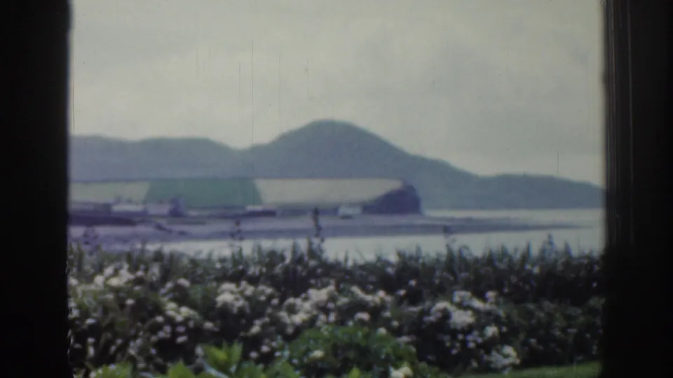 1961 Ireland home-movie view through a window, showing garden flowers, water, hills, and a large building along the shoreline.