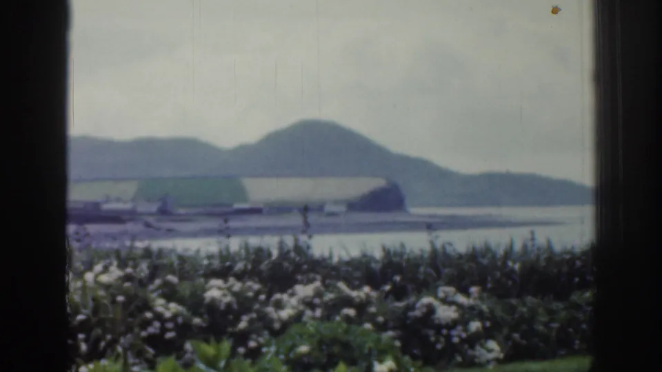 1961 Ireland home-movie frame looking past a dark window edge toward a lake or bay, green hills, and foreground flowering bushes.