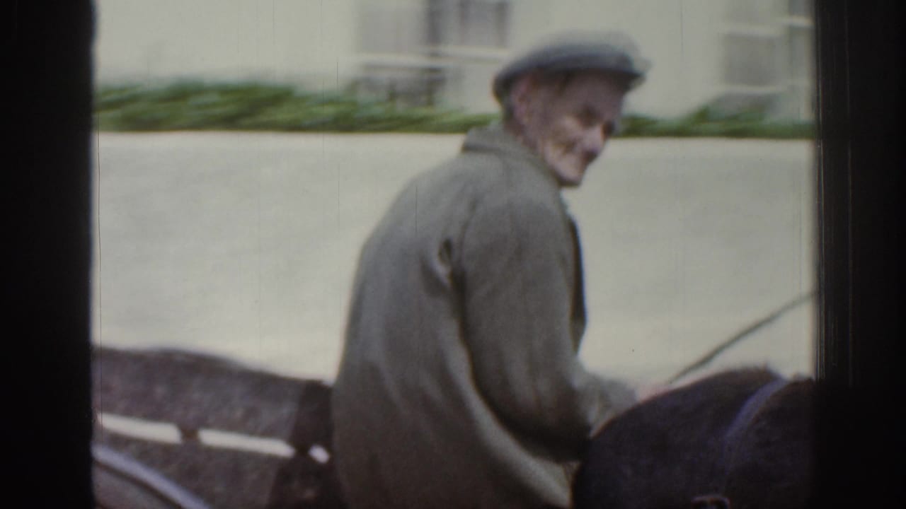 An older man wearing a flat cap and jacket rides a mule-drawn cart through a quiet Irish town street in the 1961 8mm footage.