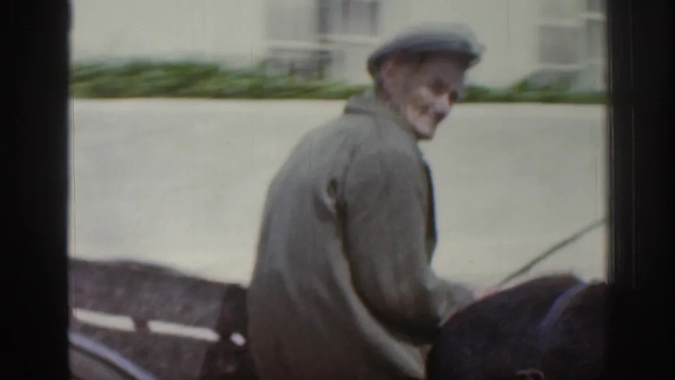 An older man wearing a flat cap and jacket rides a mule-drawn cart through a quiet Irish town street in the 1961 8mm footage.