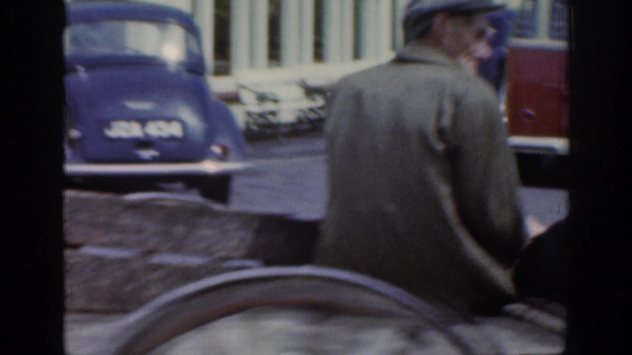 The man on the cart is seen from behind as the home-movie camera catches older parked cars and town buildings along the street.