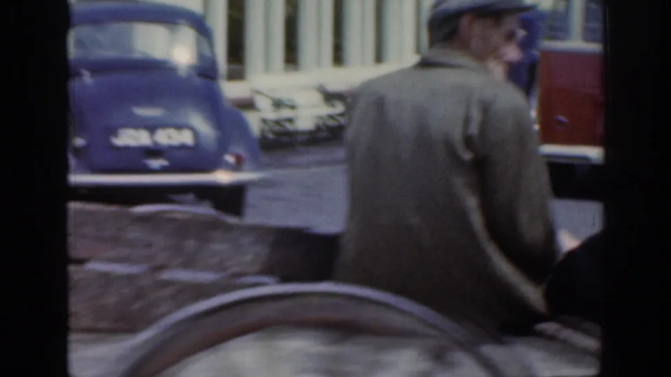 The man on the cart is seen from behind as the home-movie camera catches older parked cars and town buildings along the street.