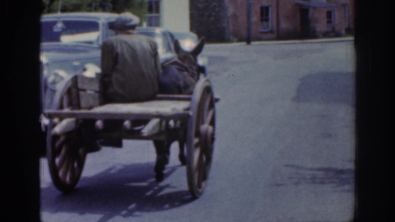 The mule-drawn cart moves away from the camera down a paved Irish town street, with the rider seated on the wooden cart and buildings nearby.