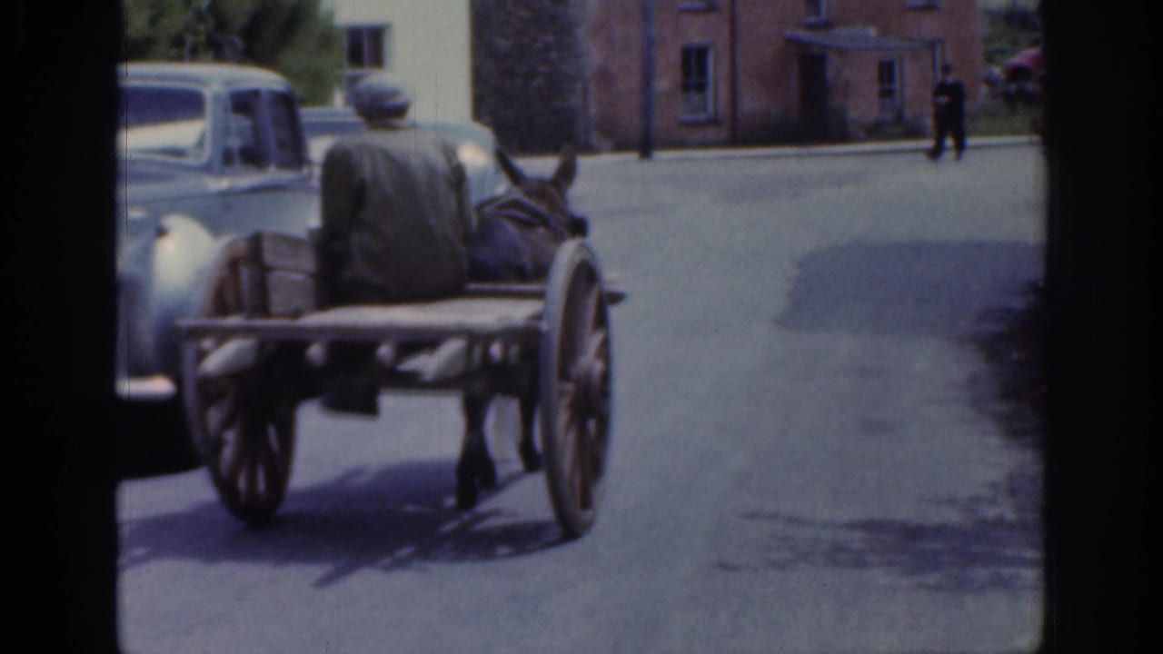 1961 Ireland home-movie frame showing a horse cart with a seated driver passing a parked car on a quiet village street.