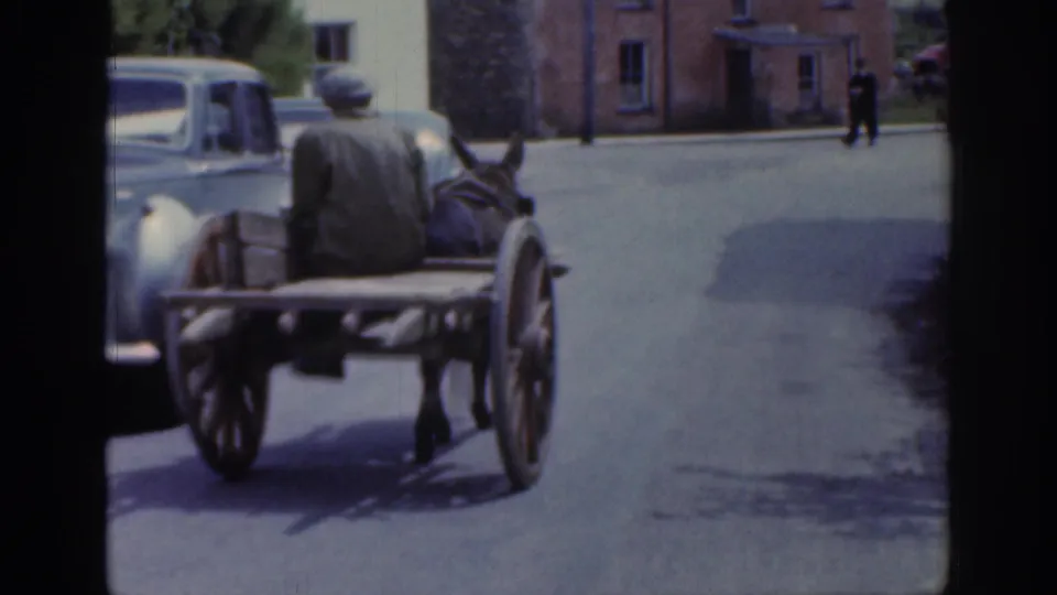 1961 Ireland home-movie frame showing a horse cart with a seated driver passing a parked car on a quiet village street.