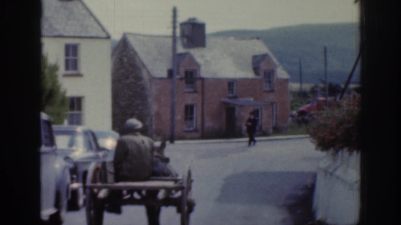 1961 Ireland frame with the horse cart receding along a sloped village road past houses and a parked car.