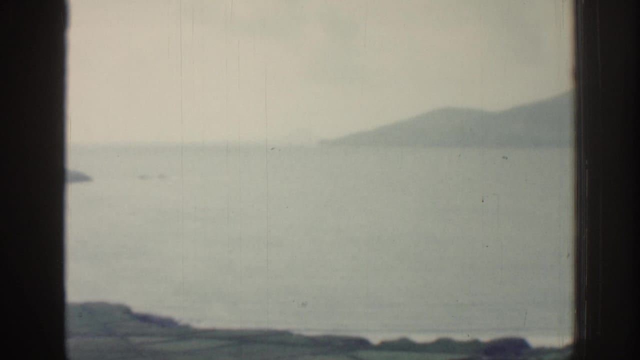 Misty Irish coastal view from the same 1961 home movie, with calm sea, low land, and distant mountains.