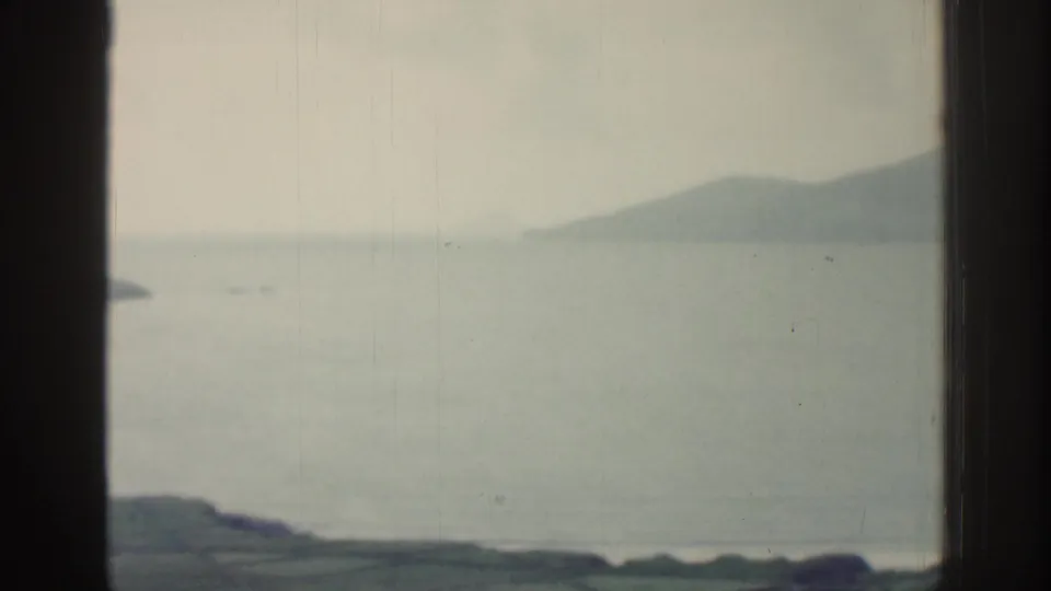 Misty Irish coastal view from the same 1961 home movie, with calm sea, low land, and distant mountains.