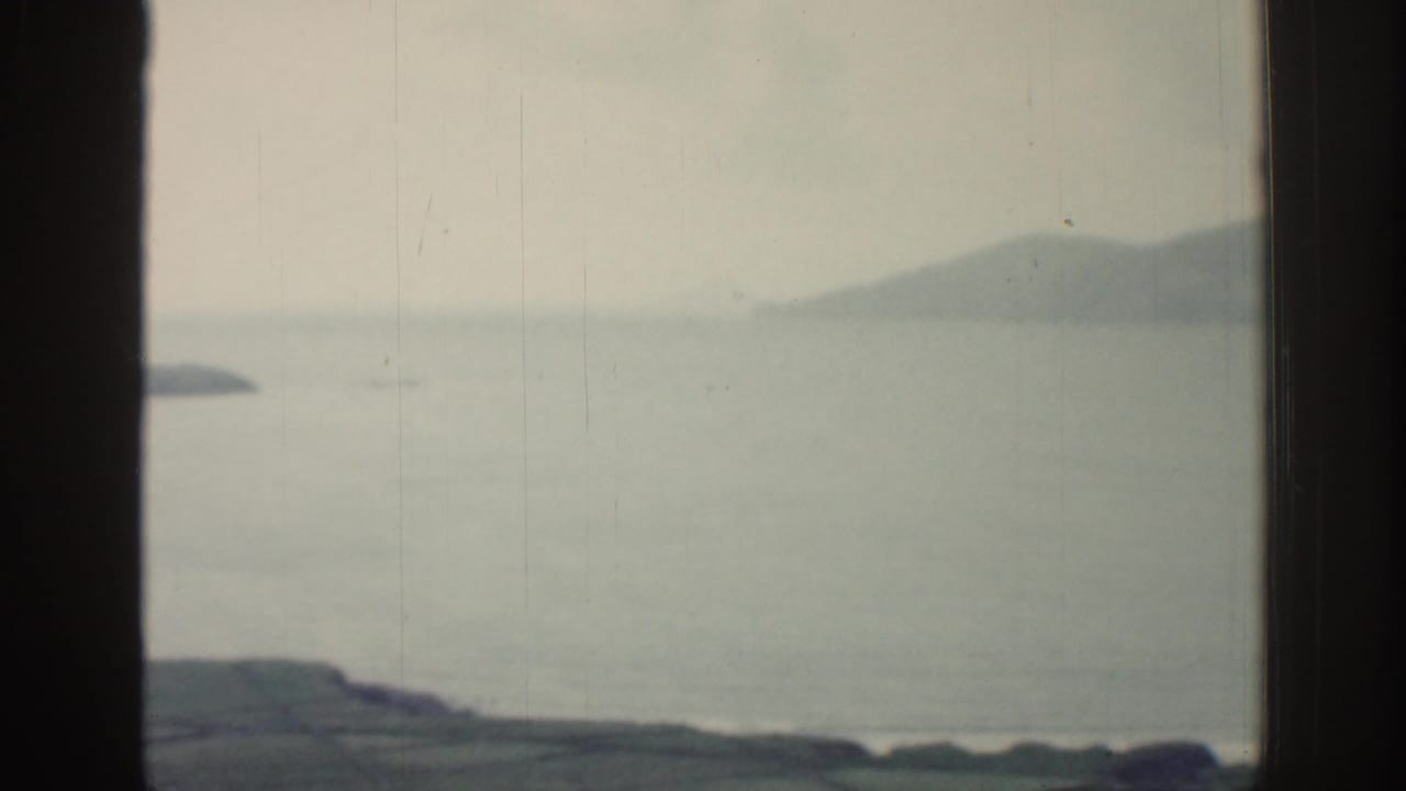 1961 Ireland coastal frame showing pale water, a dark foreground shoreline, and soft distant mountains.